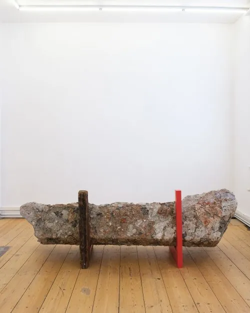 Large rough stone slab displayed horizontally, supported by a dark wooden beam and a red metal bar in a minimal gallery space.